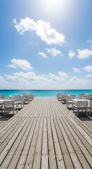 Fototapeta premium Perspective view of a wooden pier leading to the turquoise ocean with tables and chairs set up for a beachside dining experience under a bright sunny sky with fluffy clouds.