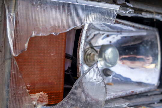 A car headlight is broken with a damaged lens and exposed wires, located in a parking area on a cloudy day needing repairs