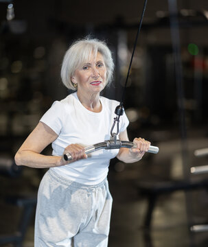 Focused elderly woman training on cable machine, performing triceps pushdowns during regular workout in gym, maintaining active and healthy lifestyle