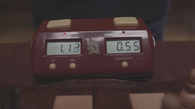 During a game of chess, players' hands hover over a digital timer. A detailed shot then shows a finger pressing the timer button to stop the countdown clock.