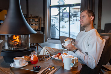 Fototapeta premium A guest in a light sweater sips coffee by a modern fireplace in a Dolomites boutique hotel, with croissants, berries, cured meats, and snow beyond large glass doors.