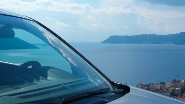 Car parked overlooking a coastal town and sea. Detail of a car hood and windshield reflecting the blue sky, parked at a scenic viewpoint with a stunning panorama of the sea, a distant island