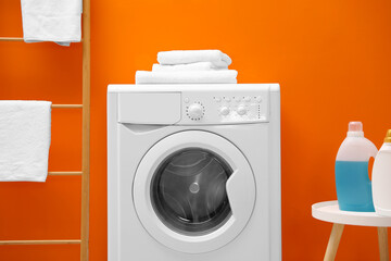 Washing machine, stack of towels and detergents near orange wall indoors