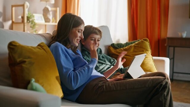 Mother son greeting tablet starting video call sitting comfortable home sofa. 