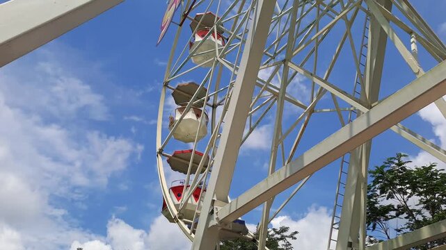 BATU, INDONESIA - DECEMBER 6, 2025: Handheld footage of a colorful Ferris wheel at a crowded amusement park in Batu, with a bright blue sky and clouds as visitors line up for the ride on a holiday.