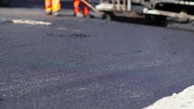 Asphalt paving machine laying fresh asphalt while workers use rakes to level the surface, and a road roller passes during street paving work.