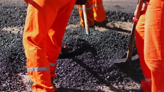 Close up of road construction workers using shovels to spread fresh asphalt during street paving and repair work.