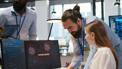 Young inexperienced developer in office requests help from team to fix hacking attempt on corporate network. Intern assisted by colleagues to investigate unauthorized access alerts, camera A © DC Studio