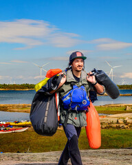 A man carrying multiple dry bags and wearing a PFD walks across a rocky terrain with wind turbines...