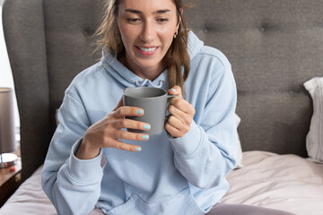 Woman sitting on bed wearing light blue hoodie holding gray mug by tufted headboard and lamp