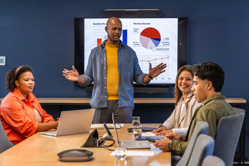 Diverse team watching standing presenter gesturing at charts on screen in meeting room with laptops