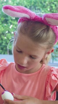 Vertical video: Daylight nudging child in pink bunny ears painting egg purple for Easter with brush