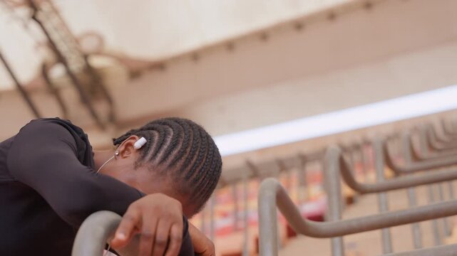 Woman resting head on metal railing, braids falling forward with earphones visible, fatigue and bowed posture suggest postpractice recovery amid empty stands and industrial beams, intimate scene