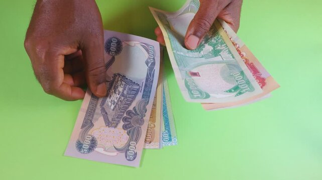 Hands carefully arrange a series of Iraqi Dinar banknotes from lowest to highest denomination on a bright green background. The video highlights the diverse colors and patterns of the currency.