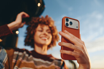 A smiling woman wearing a rainbow striped sweater takes a selfie with a smartphone outdoors during...