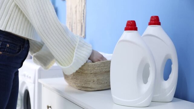 Woman putting wicker basket with clean towels onto washing machine and taking white ones near bottles of detergents and blue wall at home, closeup