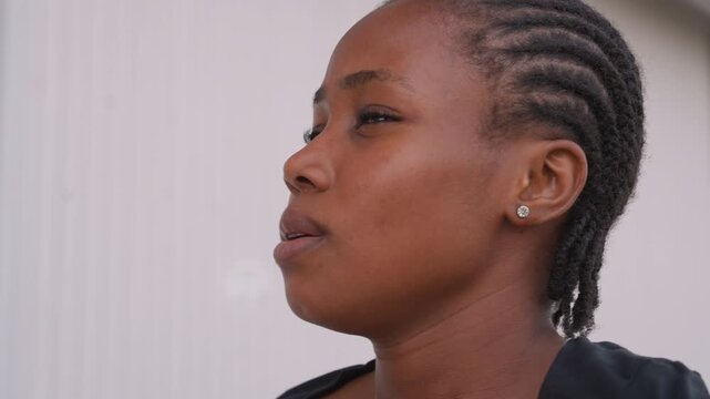 Close profile black woman with braids against pale wall, soft natural light highlighting cheekbone, contemplative gaze, subtle earring detail, minimalist urban portrait conveying quiet reflection.