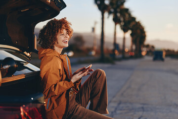 Fototapeta premium Young woman with curly hair wearing rainbow sweater sits on car trunk smiling and holding smartphone outdoors on sunny day with palm trees and road in background.