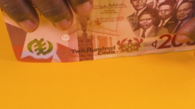 Hands carefully stack various denominations of colorful Ghana Cedi banknotes on a yellow background. The video highlights the 200 Cedi note, featuring Ghana's "Big Six" historical leaders.
