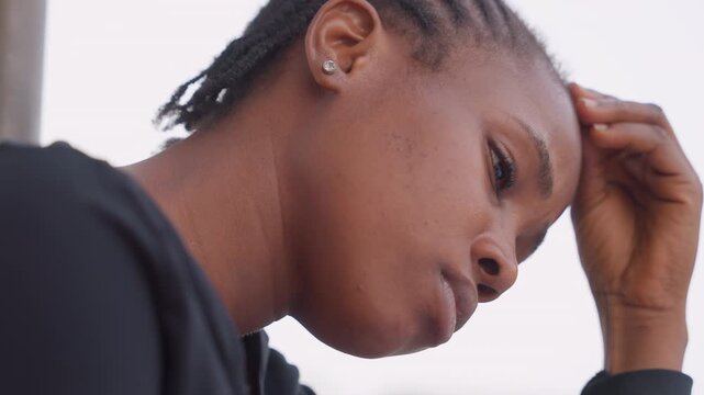 Black girl resting forehead with hand, eyes downcast and thoughtful, student pausing outdoors with cornrows and earring, quiet tension and waiting vibe, soft sky background, intimate profile study
