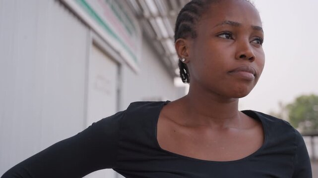 Pensive black student standing outside storefront at dusk, braided hair and arms on hips, contemplative gaze toward street, metal awning overhead, quiet urban strip, internal decision moment captured