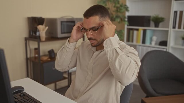 Hispanic man adjusts glasses at desk in building while seated upright and looking at screen; focus concentration.