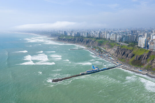 Sunny morning Miraflores Barranco Barranquito Playa Makaha beach pier aerial drone Lima Peru clear hazy blue skies residential skyscraper buildings cars traffic highway ocean view surf waves static