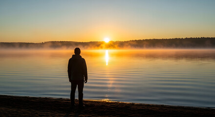 Peaceful man watching the sunrise over a misty lake at dawn, serene natural landscape