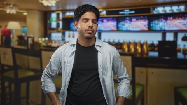 Man standing at a bar counter inside a building with arms crossed and one hand tucked into pocket, wearing casual shirt and black tee, looking toward screens; pensive.