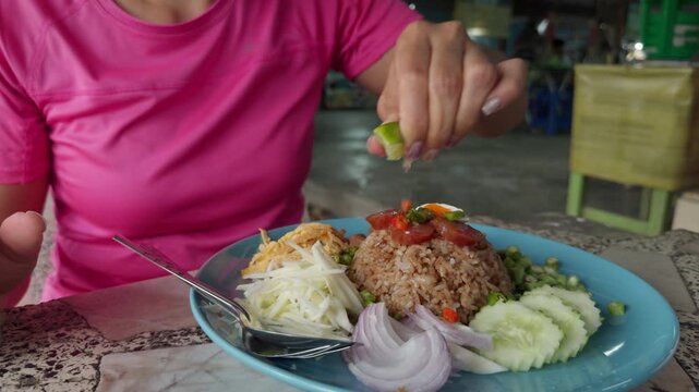 Woman squeezing fresh lime juice over traditional Thai shrimp paste rice Khao Kluk Kapi in local restaurant close up food preparation before eating