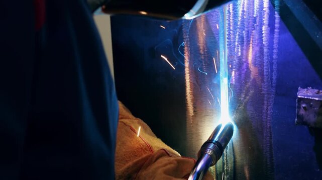Industrial welder creating vertical welding seam on steel structure with bright arc and sparks during metal fabrication process in factory workshop.
