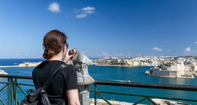 Mujer contemplando las Tres Ciudades de Malta desde un mirador con telescopio. Paisaje costero mediterr&aacute;neo y arquitectura hist&oacute;rica defensiva.