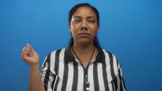 Woman referee wearing striped shirt holds whistle and points thumb in studio against blue wall; confidence.