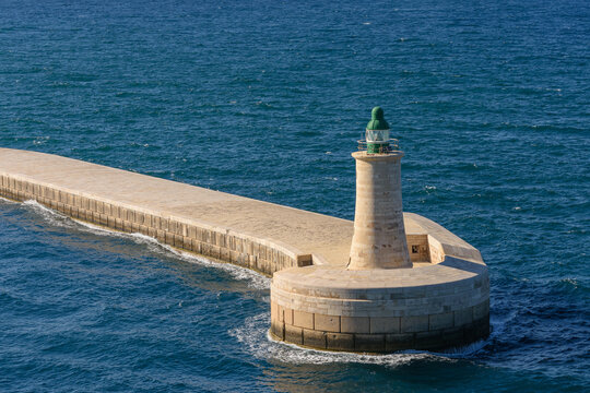 Vista en &aacute;ngulo del rompeolas y faro verde en la bah&iacute;a de La Valeta. Estructura masiva de piedra y aguas cristalinas del mar Mediterr&aacute;neo.