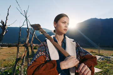 Woman with axe on shoulder on rural farm, portrait at sunrise in mountains. Outdoor worker in warm jacket, determined expression, rustic fence and field in backlit landscape. © SHOTPRIME STUDIO