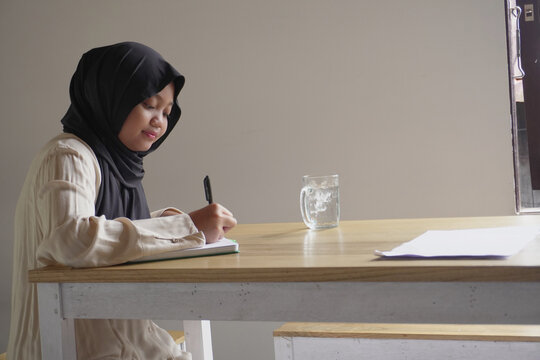 Diligent young Asian Muslim tween girl sitting alone at a cafe, writing in her personal journal or doing homework in a cozy atmosphere
