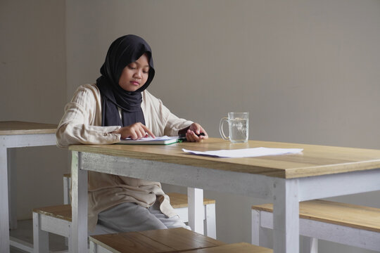 Diligent young Asian Muslim tween girl sitting alone at a cafe, writing in her personal journal or doing homework in a cozy atmosphere