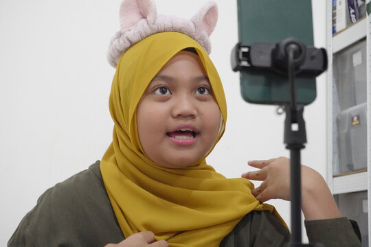 Cheerful young Asian Muslim tween girl live streaming on smartphone to sell cute furry ear headbands
