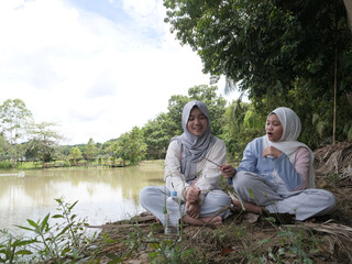 Two happy young Asian Muslim sisters sitting under a tree by the lake at sunset, chatting and laughing together