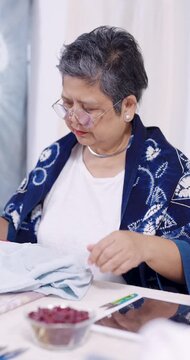 Senior Asian woman artisan picks up denim cloth, checks tablet order note, counts stitch points, then resumes removing red thread from resist stitching, preparing shibori piece for indigo dyeing.