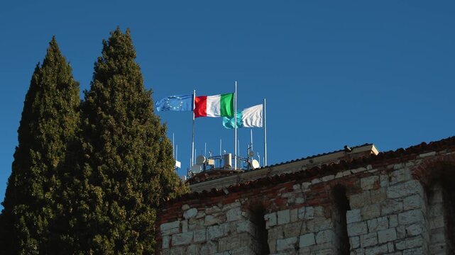 Flags of Europe, Italy and Lombardy region waving against clear blue sky at Brescia Castle, slow motion