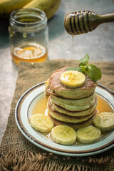 Stack of fluffy homemade banana pancakes topped with fresh slices, mint leaf, and golden honey dripping from a wooden dipper. Rustic breakfast concept on burlap background with natural sunlight. © Chayut