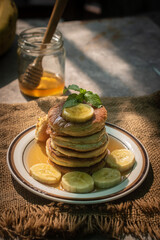 Stack of fluffy homemade banana pancakes topped with fresh slices, mint leaf, and golden honey dripping from a wooden dipper. Rustic breakfast concept on burlap background with natural sunlight. © Chayut