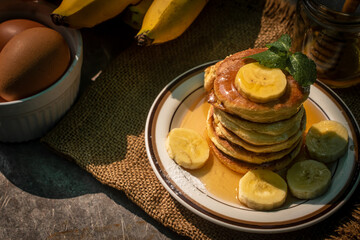 Stack of fluffy homemade banana pancakes topped with fresh slices, mint leaf, and golden honey dripping from a wooden dipper. Rustic breakfast concept on burlap background with natural sunlight. © Chayut