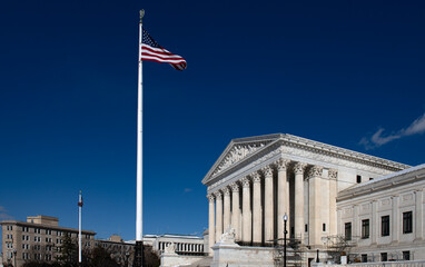 United States Supreme Court building with American flag under blue sky. Washington DC government landmark with marble columns exterior. Federal courthouse facade representing justice and law.