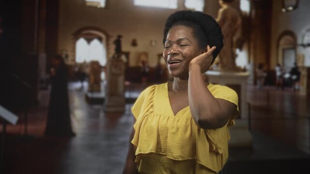 Woman touches ear with hand and looks up at a marble statue while wearing a yellow top inside a museum building; quiet curiosity.