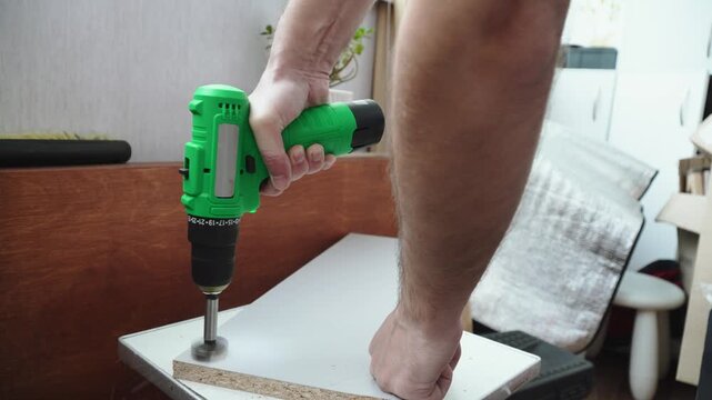 Caucasian man drilling hole for hinges in chipboard with Forstner bit and screwdriver closeup. Attaching furniture components and DIY furniture assembly and construction.