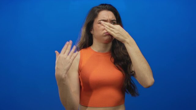 Hispanic teenager woman pinches nose in blue studio setting with visible hand gesture and wrinkled nostril; disgust aversion odor.