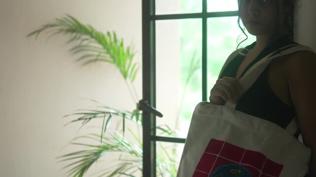 Woman holding an ecobag with a handmade design, reaching into the bag to retrieve an item, showing daily fashion and accessory use in a domestic setting