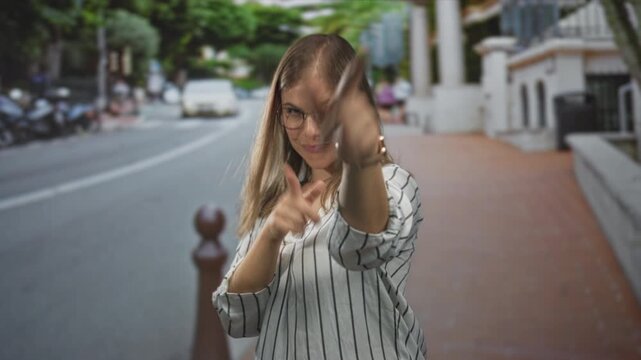 Young blonde woman wearing glasses pointing finger towards viewer on sunlit city street; playfulness.